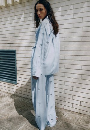 Femme aux longs cheveux bouclés portant une chemise oversize rayée bleu clair et un pantalon large assorti, debout contre un mur en briques blanches.