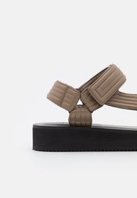 Brązowe, wyściełane sandały z grubą czarną podeszwą, z krzyżującymi się paskami i teksturowanymi akcentami. Design podkreśla komfort i stabilność.