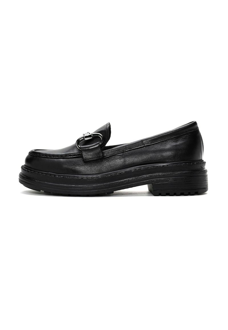 Mocasines de cuero negro con una suela plana cómoda, que presentan una hebilla decorativa de plata y detalles de costura a lo largo de los bordes.