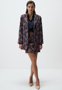 Veste de costume florale bleu marine avec une jupe assortie, présentant des motifs paisley complexes en rouge et crème. Chemise bleue brillante en dessous.
