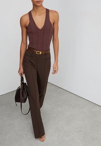 Frau trägt ärmelloses, geripptes braunes Top, hoch taillierte dunkelbraune Hose, braunen Gürtel, trägt dunkelbraune Handtasche, steht auf grauem Boden.