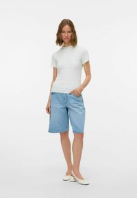 Valge tekstuuriga lühikeste varrukatega kudum, millega on kombineeritud helesinised denim bermudad. Mudel seisab ühe käega taskus, kandes heledaid jalatseid.