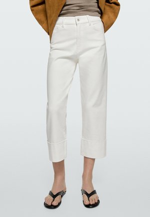 Femme portant un pantalon blanc court à jambes larges avec ourlets retournés, une veste marron, un top beige côtelé et des sandales noires à lanière entre les orteils.
