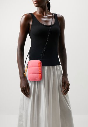 Persona vestida con camiseta sin mangas negra y falda blanca, llevando un pequeño bolso cruzado rosa con correa negra y tiradores de cremallera naranjas.