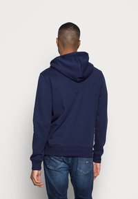 Sudadera azul marino con capucha ajustable, puños y cintura de canalé; hecha de tela suave; mostrada desde la parte de atrás, combinada con jeans azules.