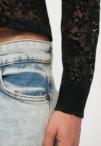 Close-up of a person wearing light blue acid-wash jeans and a black long-sleeve lace top with floral patterns.
