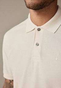 Kerman polo-paita klassisella kauluksella, kahden napin napitus ja hienovarainen logo-merkintä. Teksturoitu kangas ja lyhyet hihat.