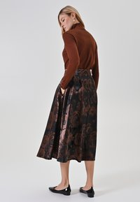 Braunes Langarmoberteil kombiniert mit einem schwarzen, knielangen Rock, der ein florales Muster in Bronze-Tönen aufweist. Das Model trägt schwarze flache Schuhe.