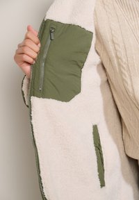 Ruka drží otvorenú béžovú fleecovú bundu s olivovozelenou zipsovou vnútornou vreckom a kontrastnými detailmi tkaniny.