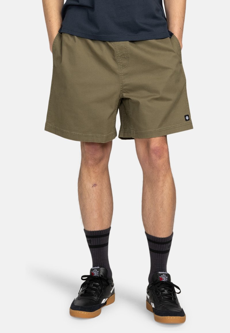 Olivgrüne Shorts mit geradem Schnitt, ausgestattet mit zwei Seitentaschen, tailliertem Saum und einem kleinen Markenetikett am linken Bein. Getragen mit schwarzen Sneakern und Socken.