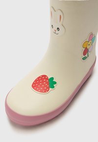 Stivale da pioggia in gomma color crema, con una suola rosa. Decorato con la faccia di un coniglio, un fiore colorato e un adesivo di fragola rossa.