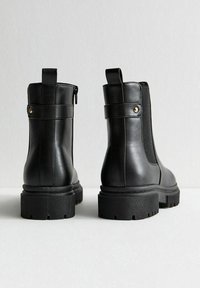 Botas de cuero negro hasta el tobillo con suela gruesa, paneles laterales elásticos y una tira de agarre en la parte posterior. Cuentan con un cierre de cremallera y herrajes decorativos.