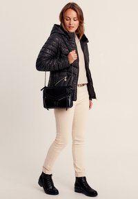 Doudoune noire avec capuche, poches zippées, pull beige, pantalon clair et bottes noires à lacets. Tient un sac à main noir.