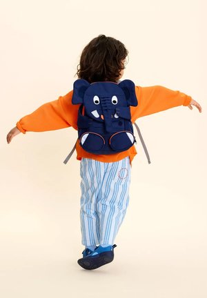 Child with curly hair wearing an orange sweater, blue striped pants, blue shoes, and a navy elephant-themed backpack, arms outstretched.