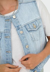Světle modrá denimová vesta s límcem, stříbrnými knoflíky a dvěma náprsními kapsami, položená na bílé tričko. Viditelná texturovaná látka.