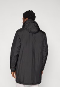 Manteau long matelassé noir avec capuche à cordon de serrage, poignets élastiques et texture lisse. Présente une coupe droite et une conception longueur genou.