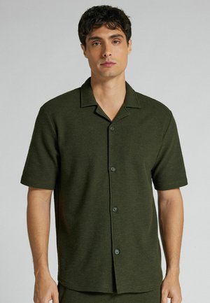 Mann mit kurzen dunklen Haaren, der ein olivgrünes kurzärmeliges Hemd mit Knöpfen vor einem einfarbigen hellgrauen Hintergrund trägt.