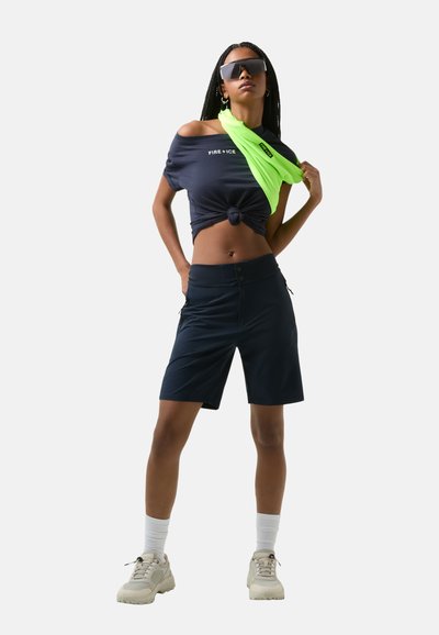 Námořnický crop top s uzlem v pase, zkombinovaný s tmavými kraťasy se zipy, doplněný jasně zeleným šátkem a béžovými teniskami.