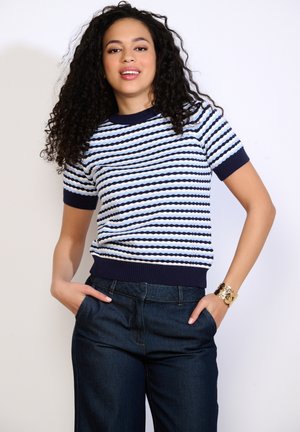 Frau mit lockigem Haar, die ein kurzärmeliges Oberteil mit marineblauem und hellblauem Muster, dunkle Jeans und ein goldfarbenes Armband trägt, mit den Händen in den Taschen.