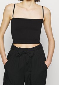 Svart ribbad crop top med tunna justerbara axelremmar, med en rak ringning och figurnära design. Bärs med hög midja svarta byxor.