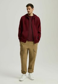 Burgundowa bluza z kapturem na zamek błyskawiczny, połączona z bordową koszulą i beżowymi spodniami cargo z bocznymi kieszeniami. Całość dopełniają białe sneakersy.