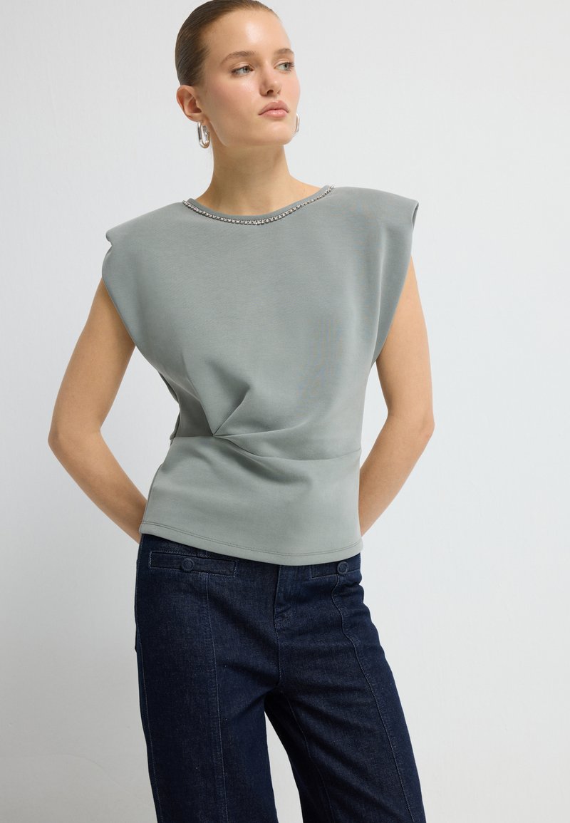 Donna che indossa un top senza maniche di colore grigio chiaro con spalline e jeans a vita alta blu scuro, in piedi contro uno sfondo chiaro e semplice.