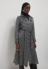 Schwarz-weißes Pünktchenkleid mit Kragen, Knopfleiste und Taillengürtel, langen Ärmeln und schwingendem Saum.