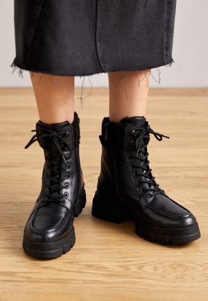 Bottines noires en cuir à lacets avec semelles épaisses portées avec une jupe noire à ourlet effiloché, debout sur un sol en bois.