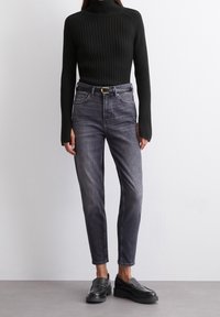 Un pull-over à col roulé noir côtelé associé à un jean gris taille haute. Présente un design ajusté et une accentuation par une ceinture argentée. Des mocassins noirs complètent le look.