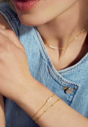 Guldhalsband och armband med skira mönster, ett prytt med pärlor. Bärs över en denimtop, vilket visar upp olika texturer och ytor.