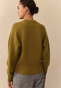 Frau mit zu einem Dutt gebundenem Haar, die einen olivgrünen Strickpullover und graue karierte Hose trägt, mit dem Rücken zu einem schlichten beigen Hintergrund.