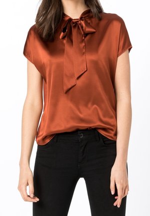 Femme portant un chemisier en satin couleur rouille avec un col noué, associé à un pantalon noir taille haute sur fond blanc.