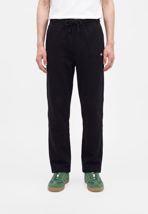 Person iført sorte snørrebukser og grønne sneakers står mod en hvid baggrund.