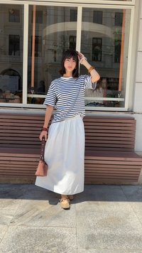 Stribet kortærmet skjorte, hvid flydende nederdel, brun taske med nitter, beige sko. Person står foran et glasmester, en træbænk er synlig.