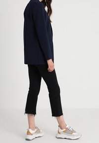 Femme portant un blazer bleu marine, un jean noir coupé avec des ourlets effilochés, et des baskets blanches avec des accents beige et gris, debout devant un fond uni.
