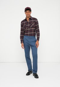Rød og marineblå rutete skjorte med lange ermer og to brystlommer, kombinert med blå bukser og sorte loafers; modell som har på seg en burgunder caps.
