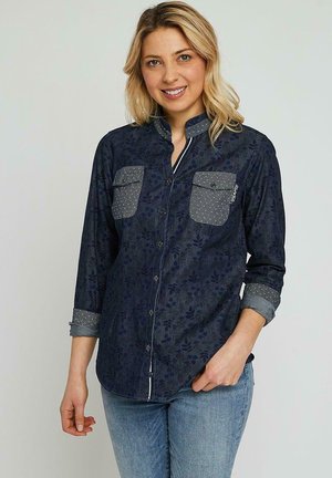 Chemise bleu marine à boutons avec motif floral, poches à carreaux contrastées et manches retroussées. Fabriquée en tissu léger avec une finition texturée.