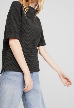 Vrouw draagt een zwart getextureerd T-shirt met korte mouwen en lichtblauwe spijkerbroek tegen een effen grijze achtergrond.