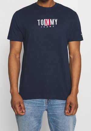 Mężczyzna nosi granatowy t-shirt Tommy Jeans z białym i czerwonym logo, w połączeniu z jasnoniebieskimi dżinsami, stojąc na jednolitym tle.