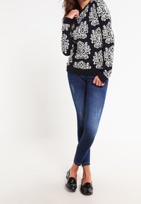 Granatowy sweter z białym kwiatowym wzorem; długie rękawy, dekolt w kształcie litery V, ściągacz na dole. Połączony z niebieskimi, dopasowanymi dżinsami i czarnymi mokasynami.