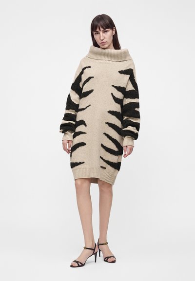 Γυναίκα που φοράει ένα μπεζ oversized πουλόβερ φόρεμα με μαύρες αφηρημένες ρίγες και μαύρα σανδάλια με ψηλό τακούνι σε απλό λευκό φόντο.