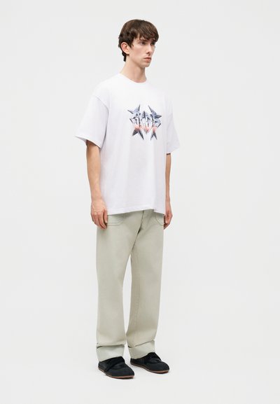 Νέος άνδρας που στέκεται φορώντας λευκό κοντομάνικο μπλουζάκι με γραφικά, ανοιχτό μπεζ φαρδιά παντελόνια και μαύρα αθλητικά παπούτσια μπροστά σε απλό ανοιχτό φόντο.