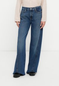 Τζιν παντελόνι με φαρδιά μπατζάκια σε σκούρο μπλε χρώμα με λεπτομέρειες ραφής, μπροστινές τσέπες και ψηλή μέση, συνδυασμένο με μαύρα επίπεδα παπούτσια.