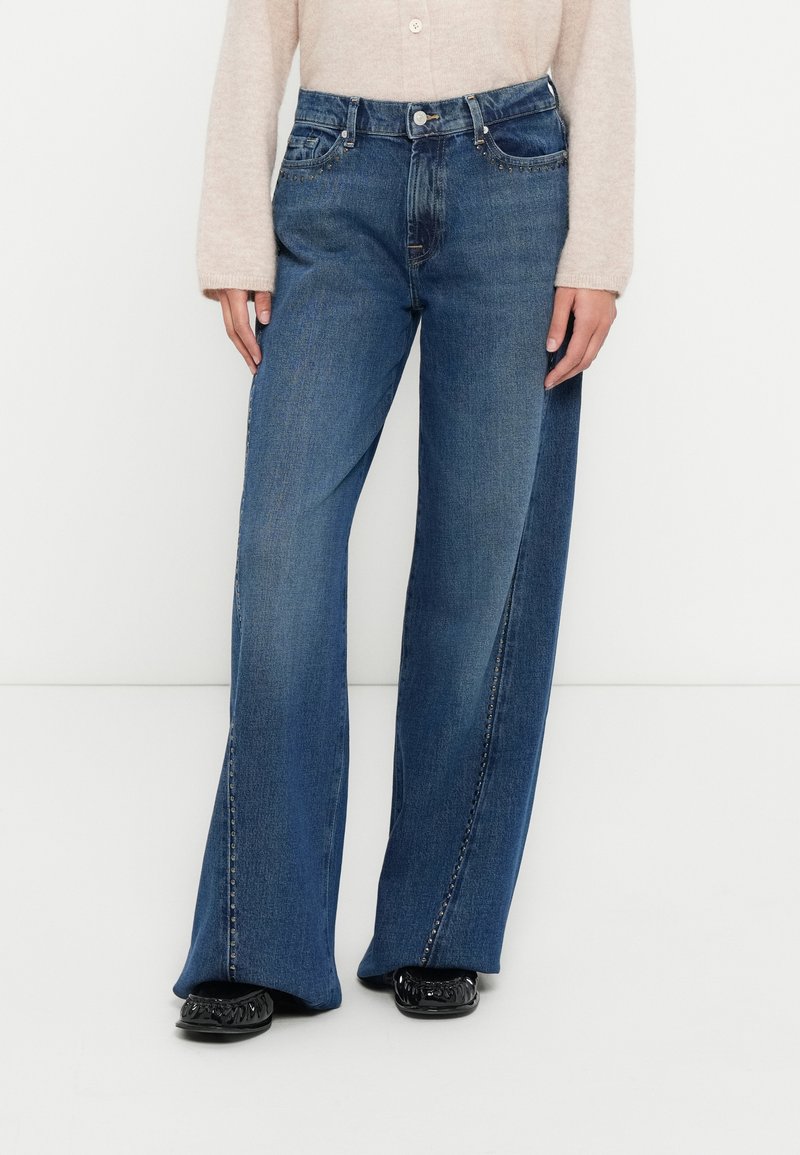 Τζιν παντελόνι με φαρδιά μπατζάκια σε σκούρο μπλε χρώμα με λεπτομέρειες ραφής, μπροστινές τσέπες και ψηλή μέση, συνδυασμένο με μαύρα επίπεδα παπούτσια.