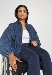 Denim jakna z sproščenim krojem, napihnjenimi rokavi in gumbi, nosena čez belo majico in v kombinaciji s temnimi jeansami. Srebrni prstani krasijo roko.