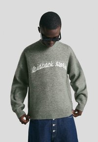 Person in einer dunklen Sonnenbrille, grauem Sweatshirt mit dem Text "laidback stories" und blauen knöpfbaren Jeans, hält den Saum des Sweatshirts.