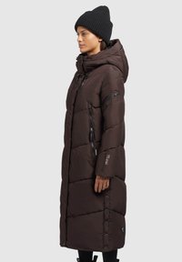 Dlouhý hnědý péřový kabát s kapucí, prošívaný design, přední zip a boční kapsy. Má rovný střih a knoflíkový uzávěr na spodním lemu.