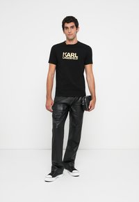 Vīrietis, ģērbies melnā Karl Lagerfeld t-kreklā, melnās ādas biksēs un melnās sneakers ar baltām zolēm, tur nelielu melnu maisiņu pret baltu fona.