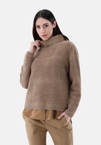 Beiger Strickpullover mit hohem Kragen, der ein strukturiertes Muster und gerippte Bündchen aufweist. Kombiniert mit einem braunen, geschichteten Hemd darunter.