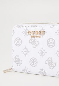 Portafoglio bianco con motivo a logo grigio impresso, hardware dorato e finitura in pelle texturizzata. Presenta il marchio GUESS sul fronte.